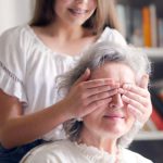 Game Library - Cheerful teenager playing with grandmother guess who game while making surprise in light living room