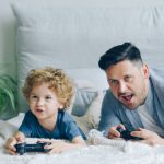 Budget Gaming - Man and His Little Son Lying on a Bed and Playing a Video Game