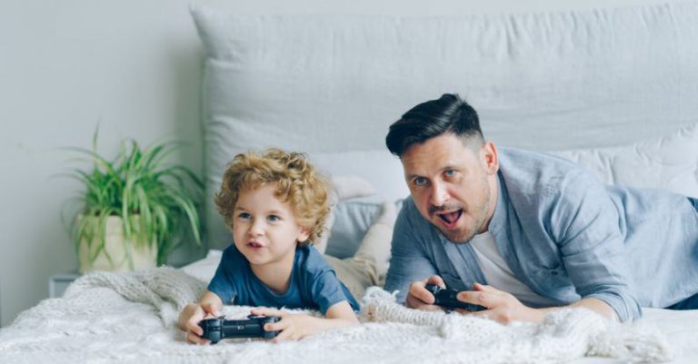 Budget Gaming - Man and His Little Son Lying on a Bed and Playing a Video Game